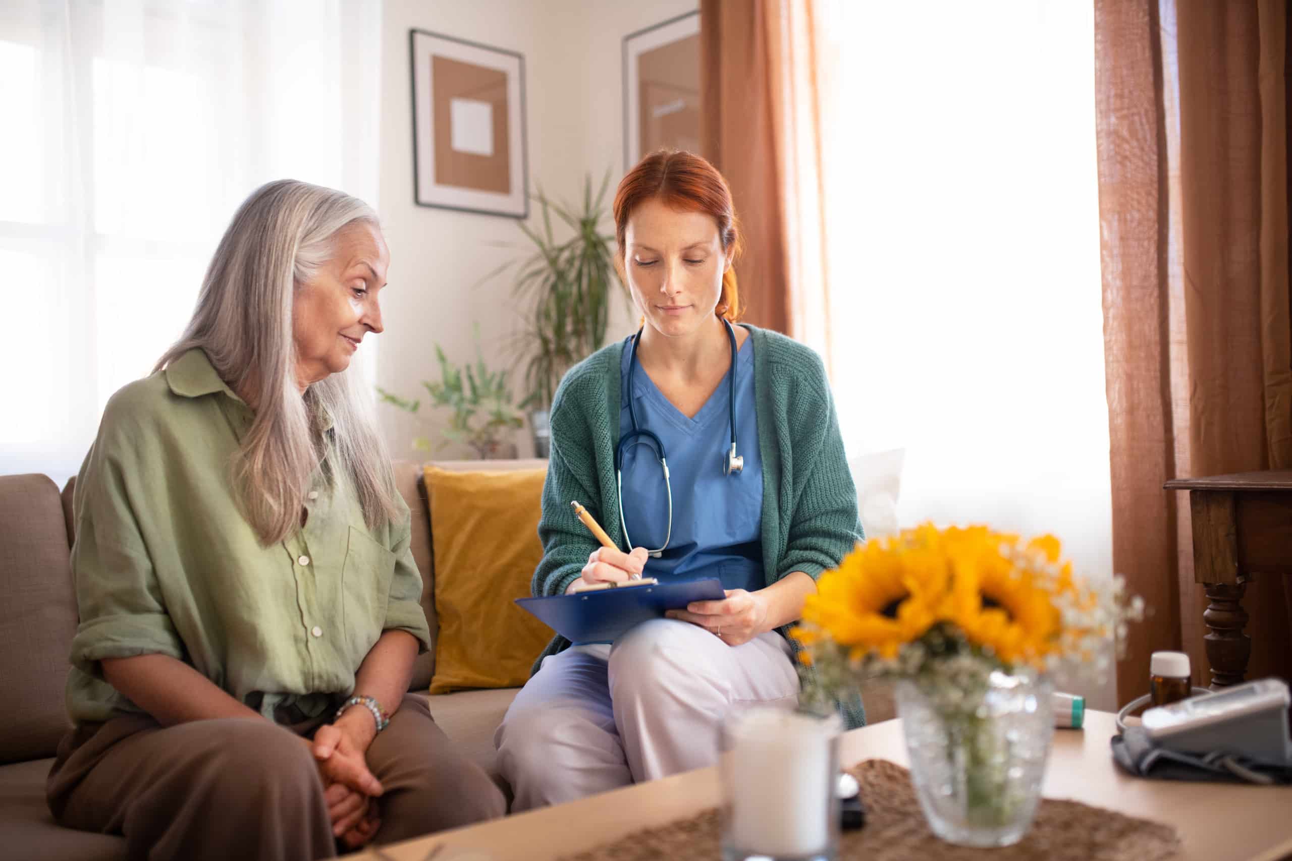 A nurse is discussing health insurance benefits with an older woman in a living room.