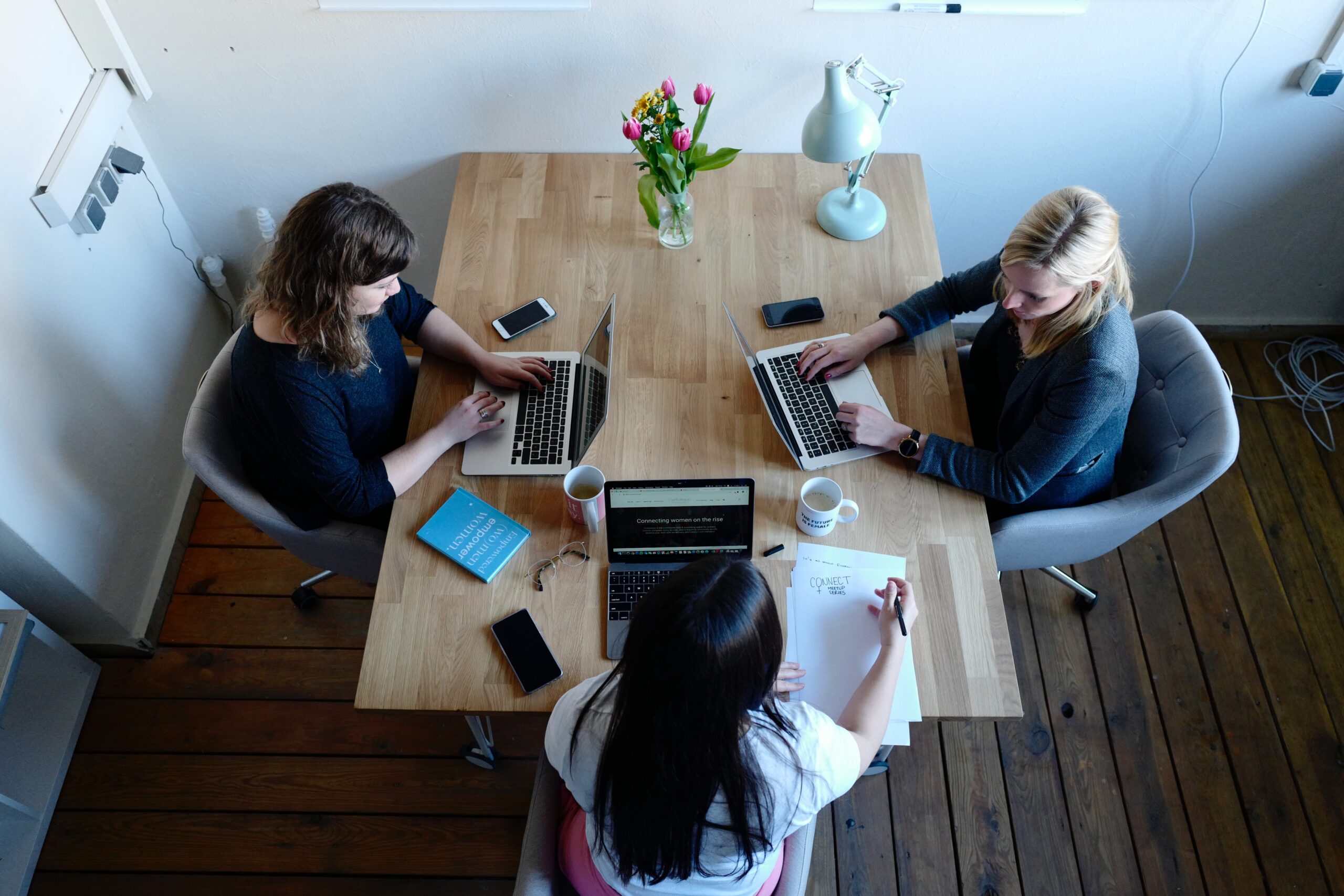 Three women working on laptops at a wooden table while discussing group health insurance options.